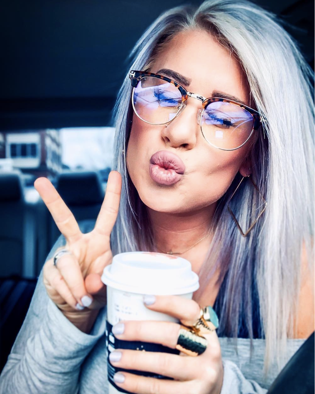 Woman with tortoise ladyboss glasses and blonde hair holding a coffee cup, making a peace sign.